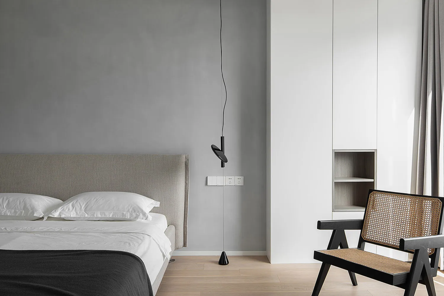 Modern minimalist bedroom featuring a bed with a gray fabric headboard, a hanging pendant light, and a woven cane chair.