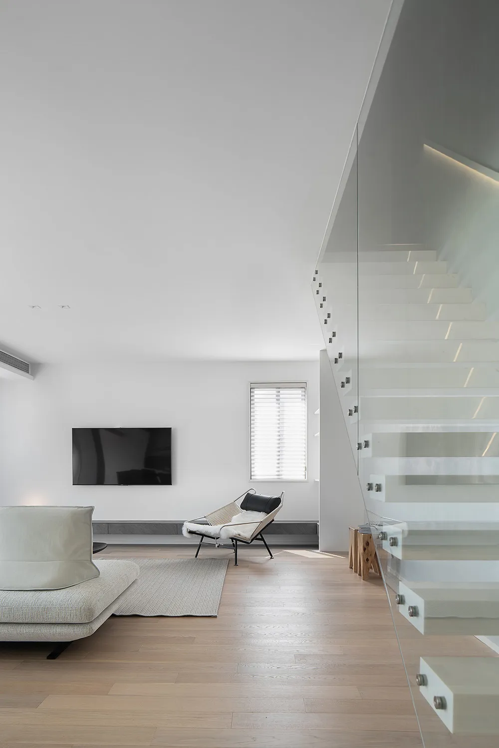 Modern minimalist living room with white furniture, wooden floors, and a glass staircase.