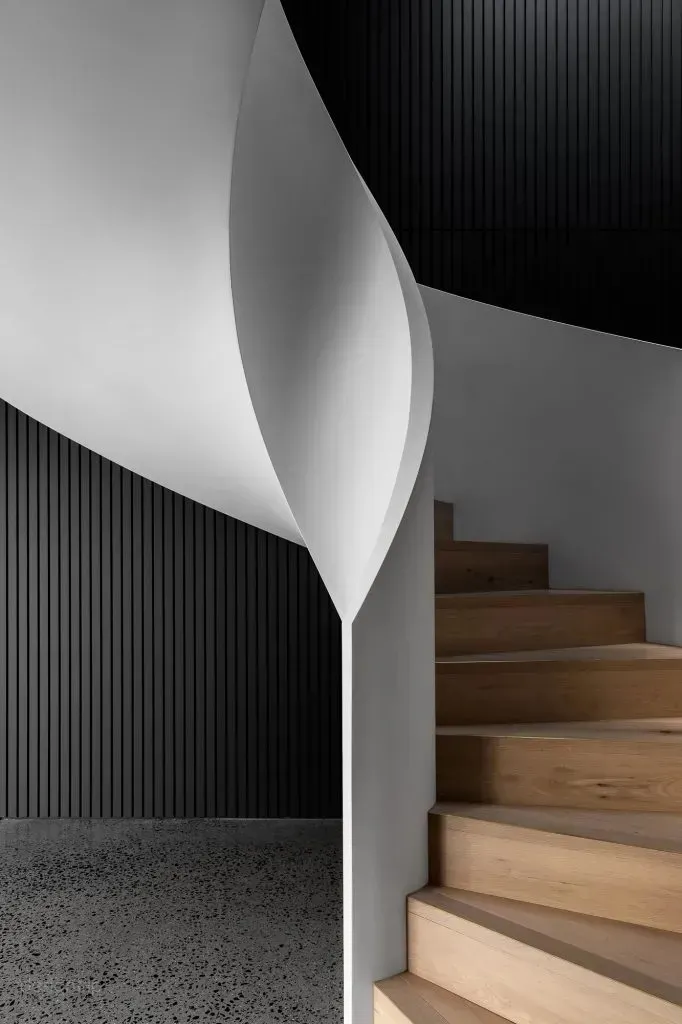 Modern minimalist staircase with wooden steps against a black and white wall, featuring sleek, curved architectural design.