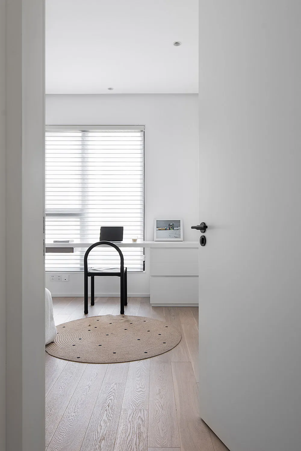 A minimalist home office with a wooden desk and black chair, featuring a round rug and a window with blinds.