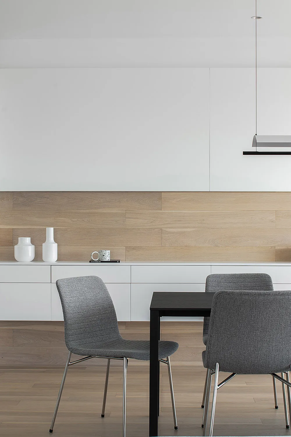 A modern dining room featuring a black table, grey upholstered chairs, and a wooden wall with white floating shelves.
