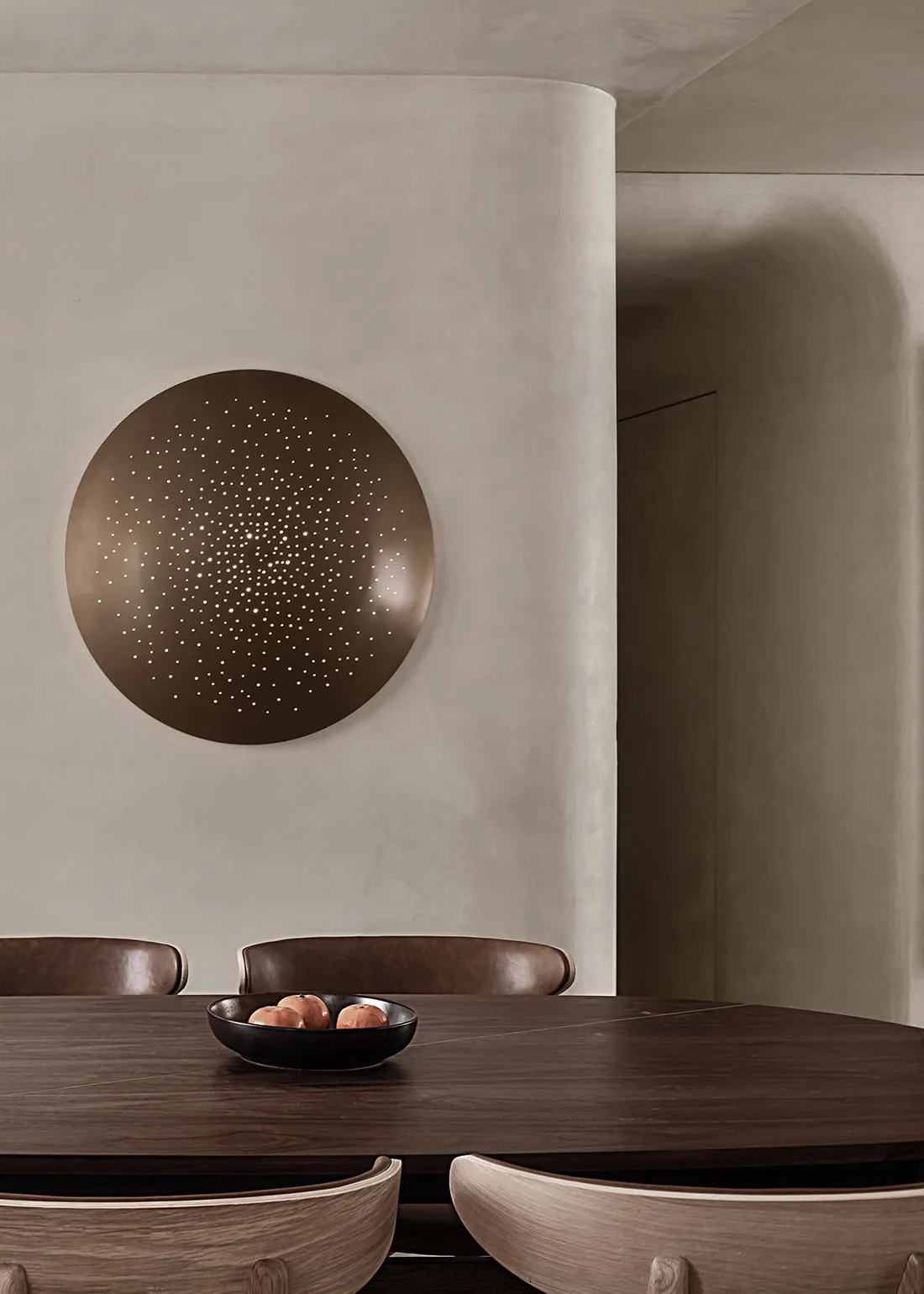 A minimalist dining room with a wooden table and chairs, featuring a round wall art piece with perforations and a bowl of app