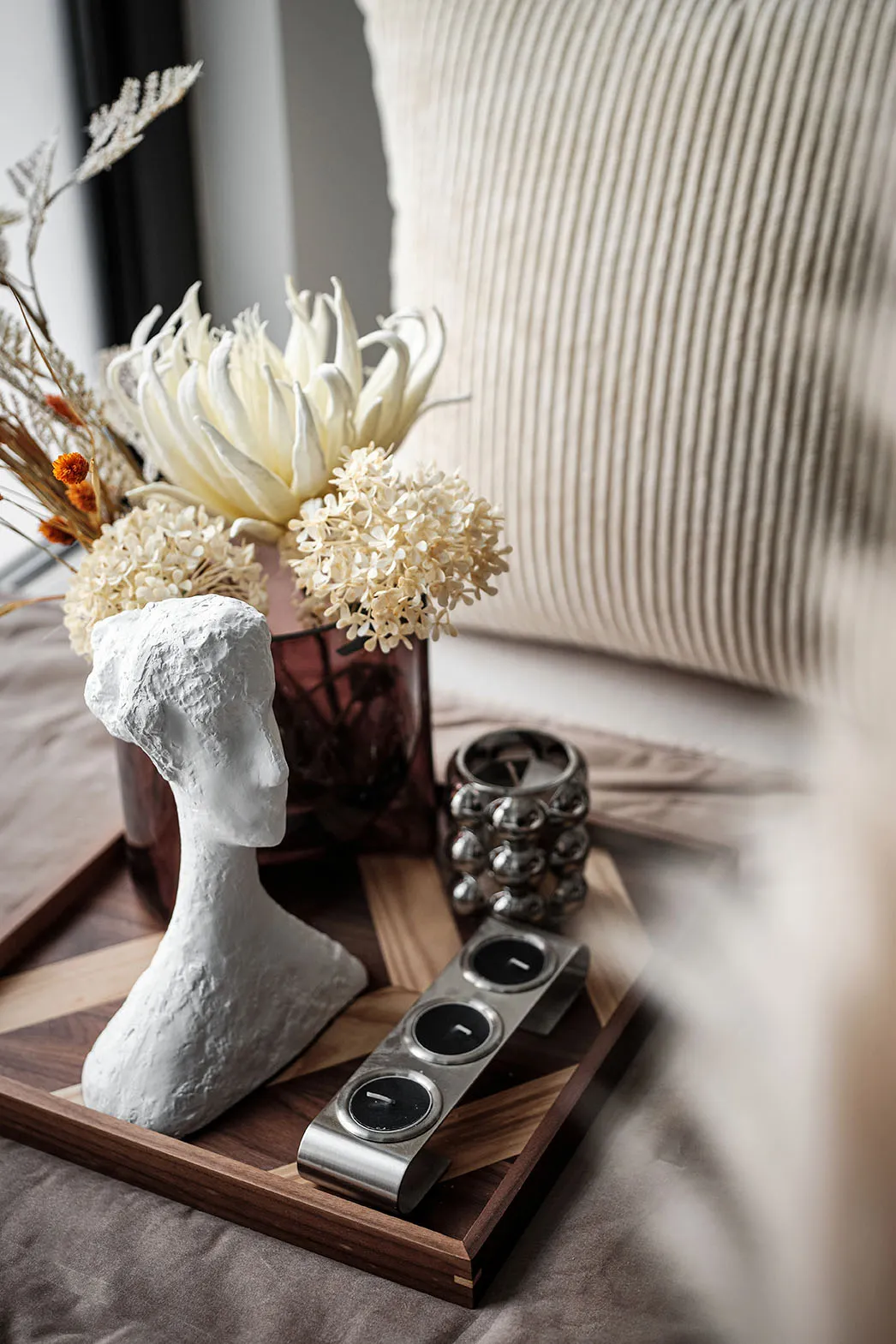 A decorative table display featuring a wooden tray, a ceramic bust sculpture, dried flowers, and metal candle holders on a be