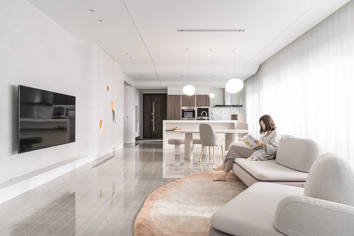 A modern interior living space with neutral tones, featuring a large sofa, a coffee table, a TV, and a dining area in the bac