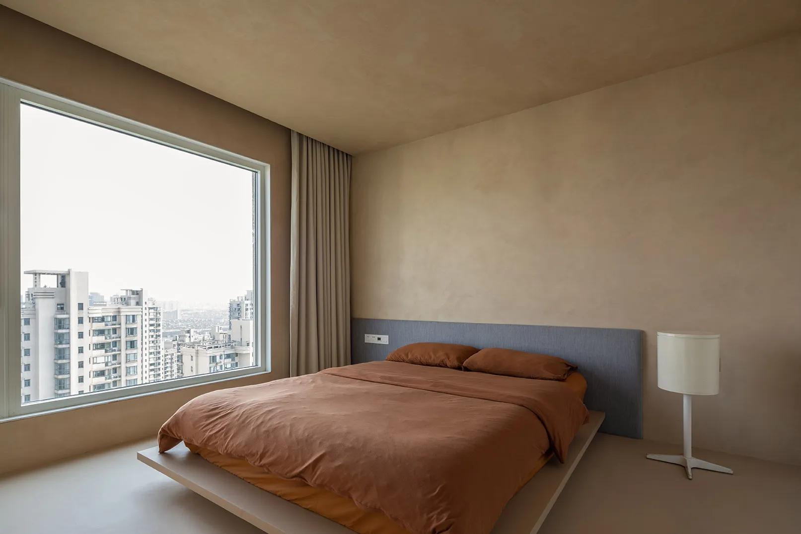 A minimalist bedroom featuring a bed with a sand-colored bedspread and a simple bedside lamp.