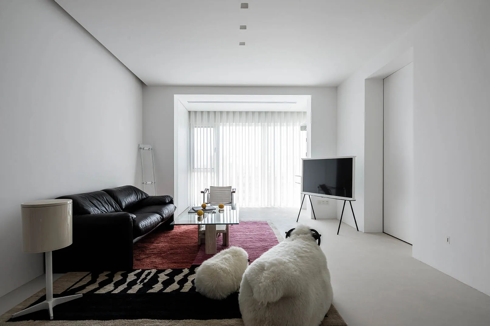 A minimalist living room with concrete walls and floor, featuring a black leather sofa, glass coffee table, and a television