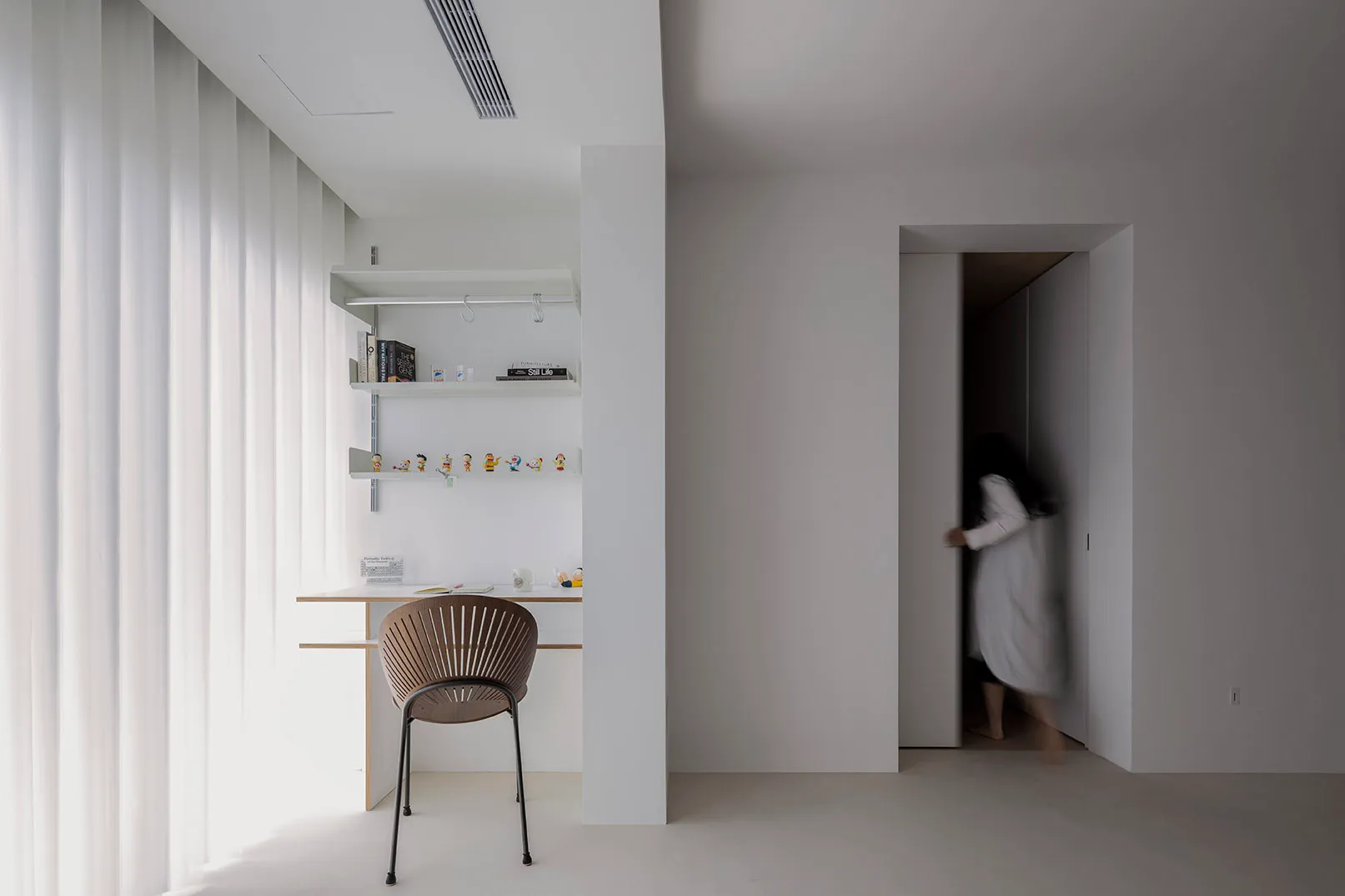 A minimalist residential interior featuring a white concrete wall, white floor, and a modern desk with shelves and a chair.