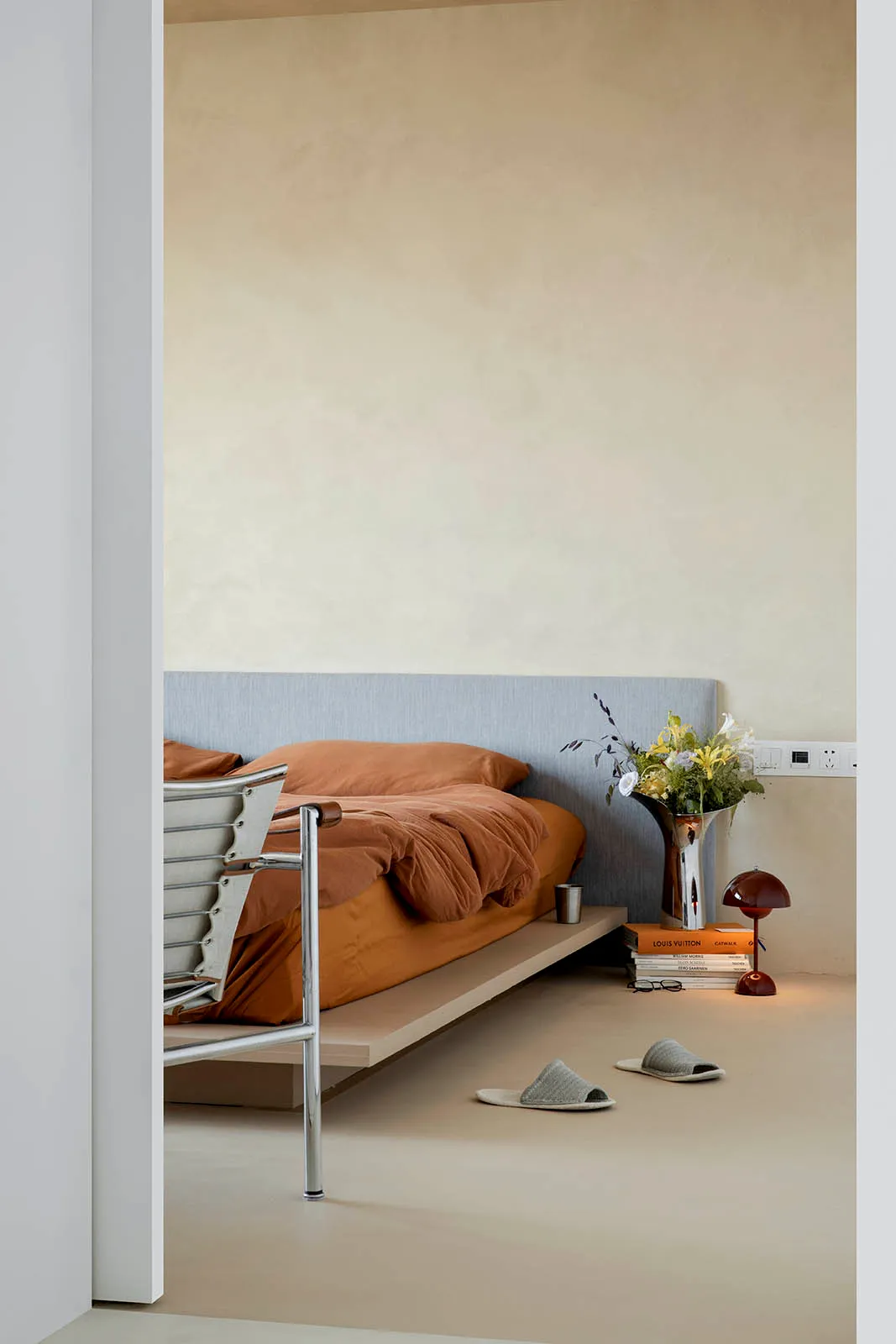A minimalist bedroom with neutral tones, brown blanket, white slippers, and flowers on the floor