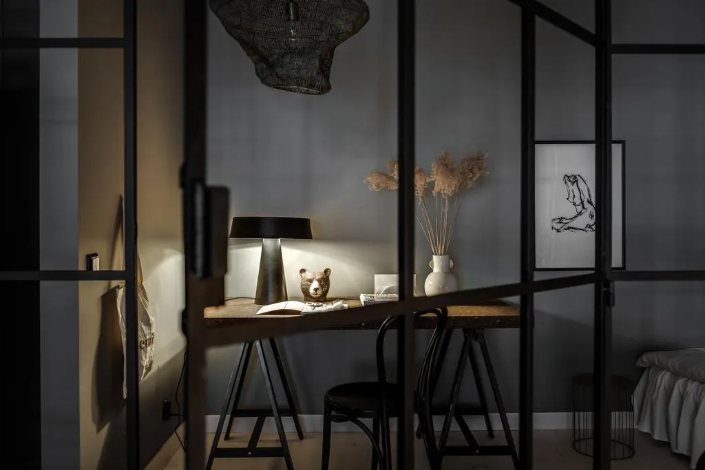 A minimalist home office space featuring a wooden desk with a black lamp and a bear figurine, positioned behind a glass parti