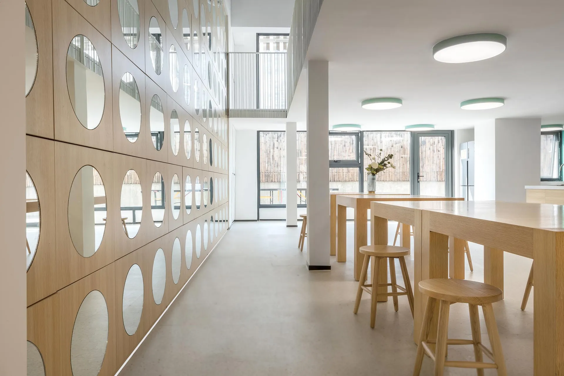 Modern minimalist restaurant interior with wood tables and stools, round mirrors, and large windows.
