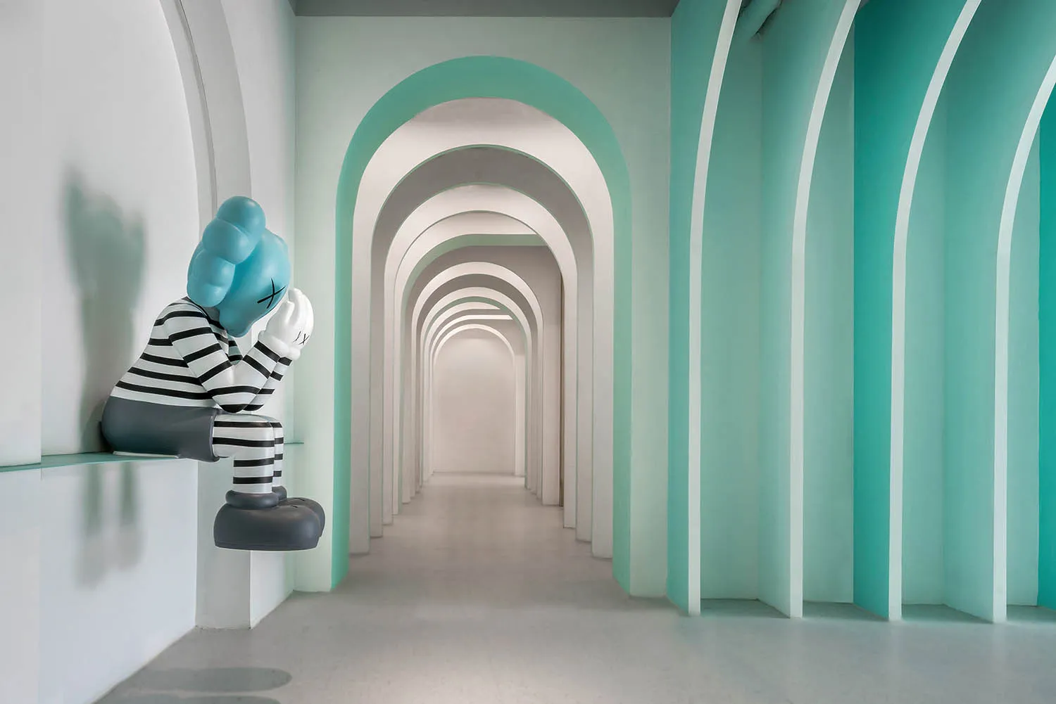 Blue sculpture with a striped black and white body sitting in a minimalist corridor with white and aqua walls.