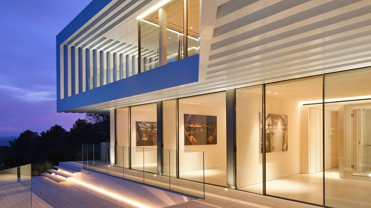 A modern residential exterior featuring glass walls and a striped glass facade at dusk.