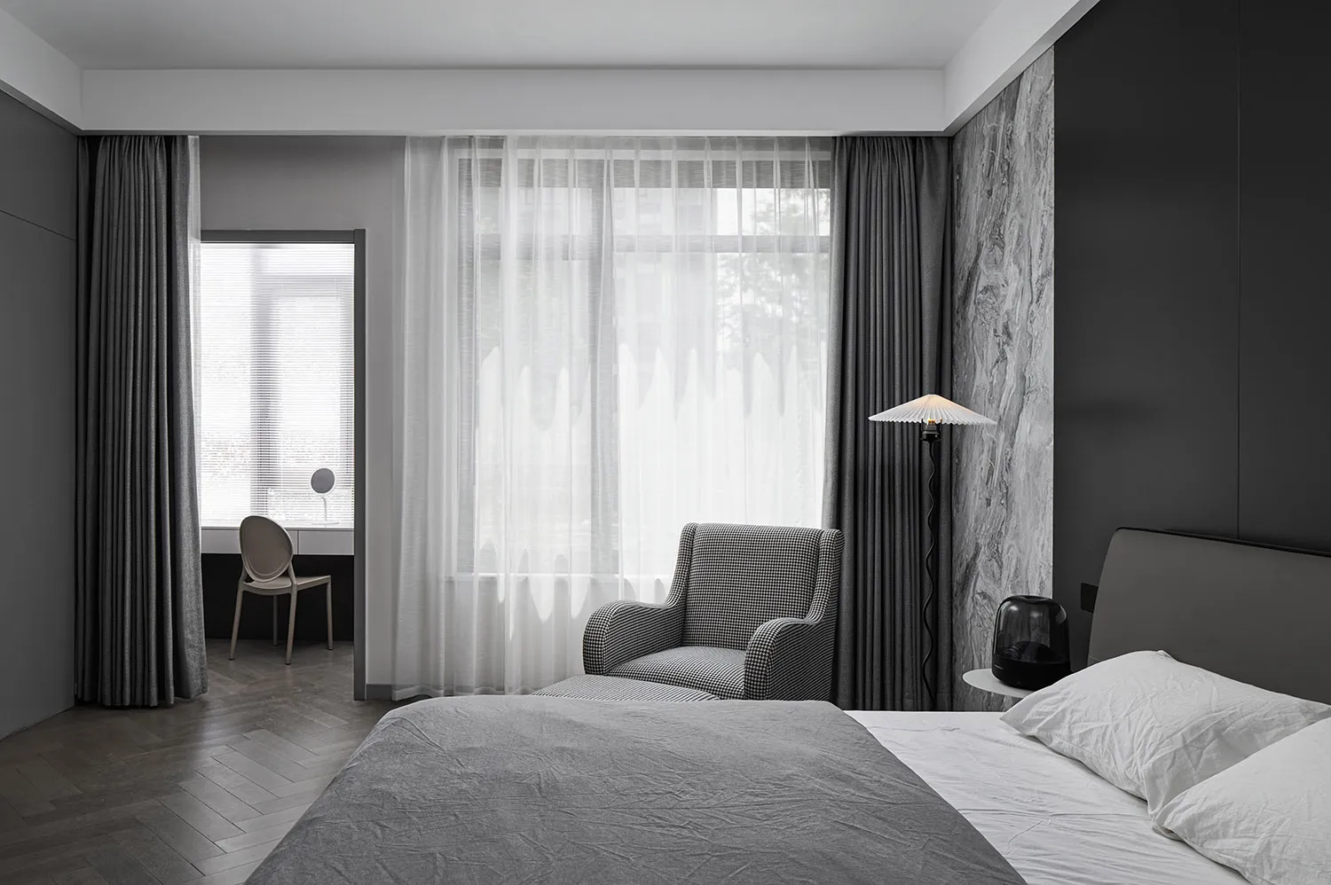 A modern minimalist bedroom with light gray walls, a bed covered in light gray bedding, a patterned gray armchair, and sheer