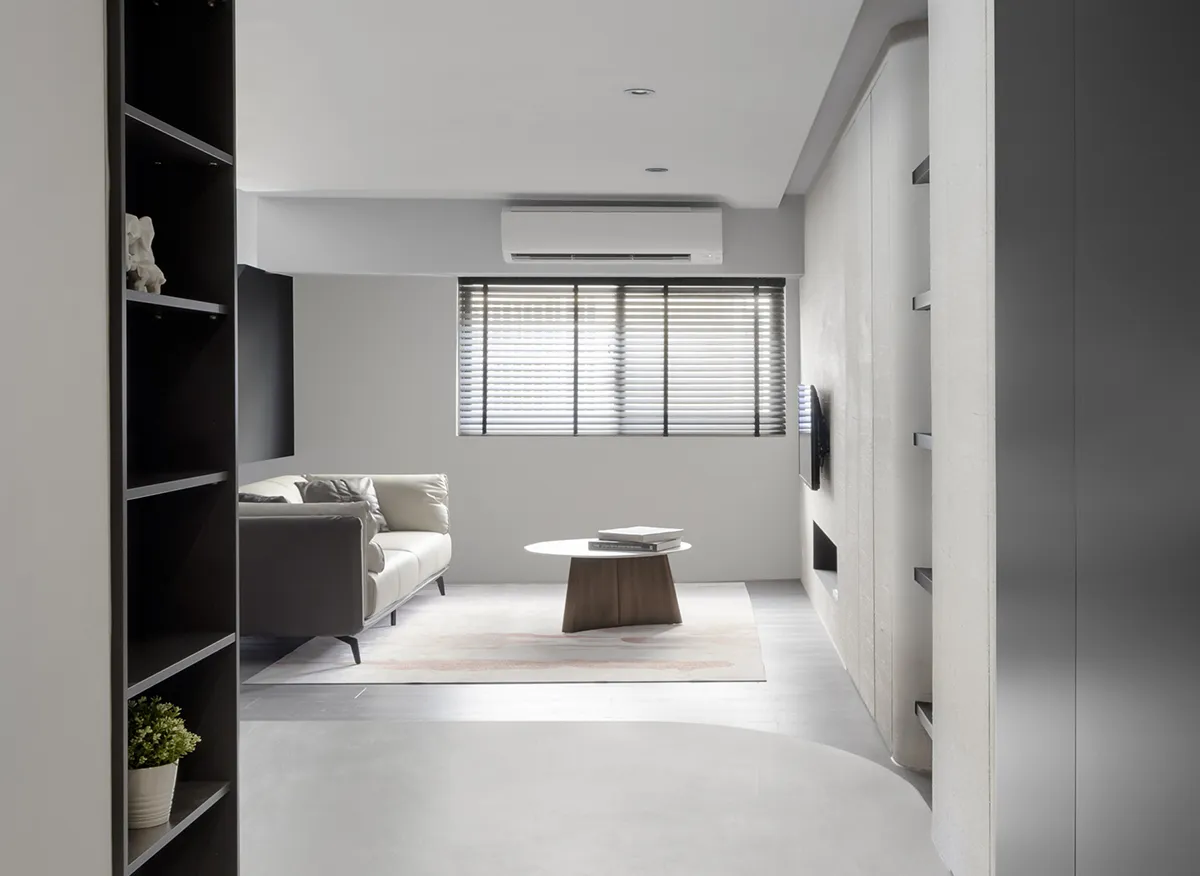 A modern minimalist living room with concrete flooring, a leather sofa, wooden coffee table, and a bookshelf with a plant.