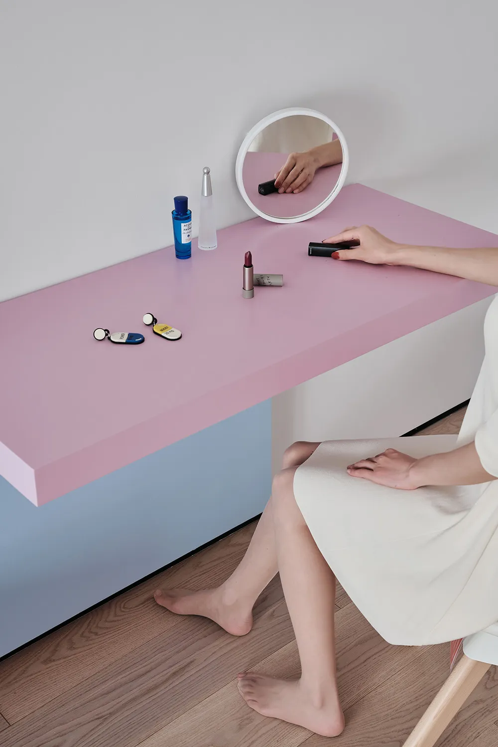 A minimalist beauty counter with a pink surface, a small round mirror, and various cosmetic items and keys.