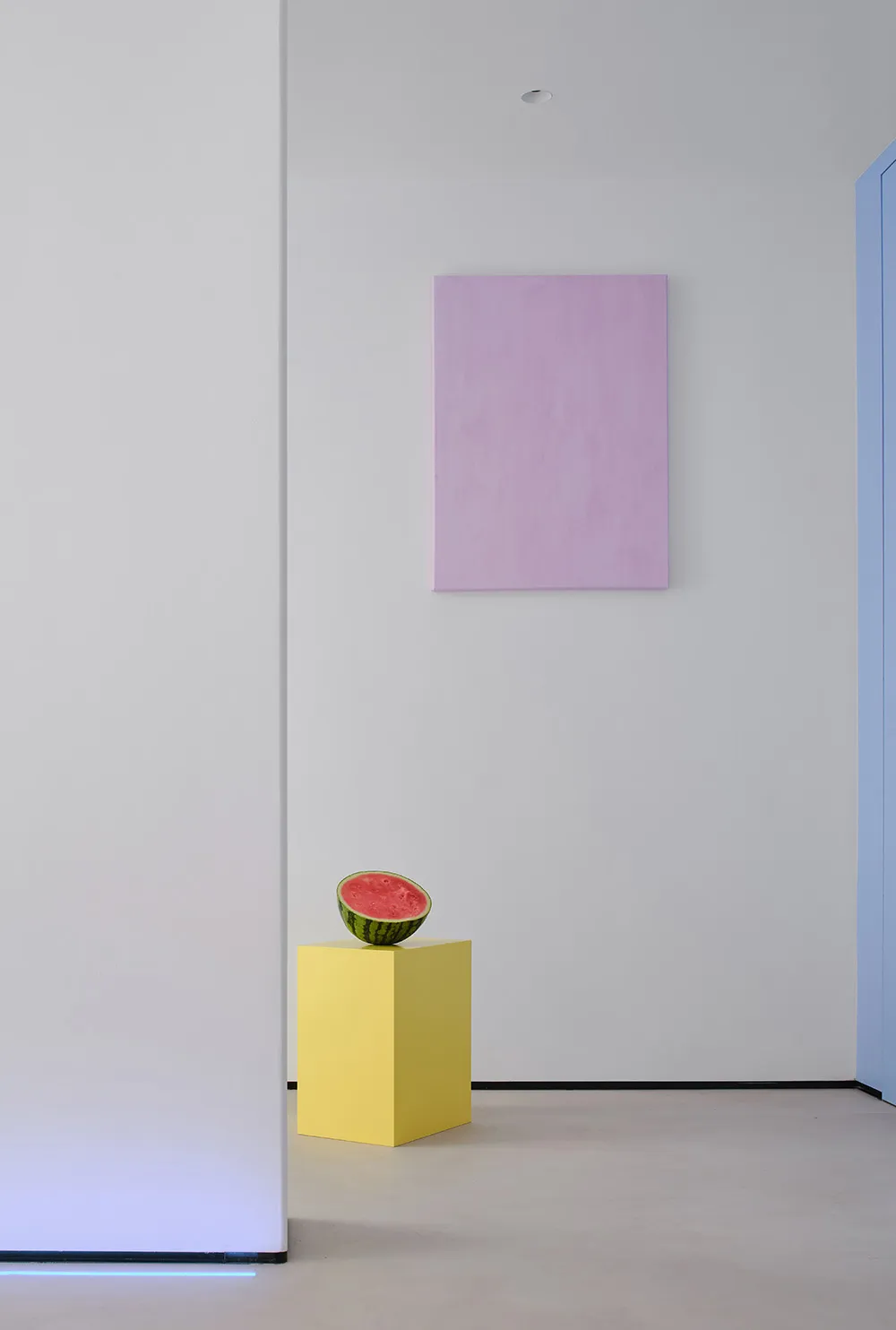 A minimalist white room with a yellow pedestal holding a half of watermelon.
