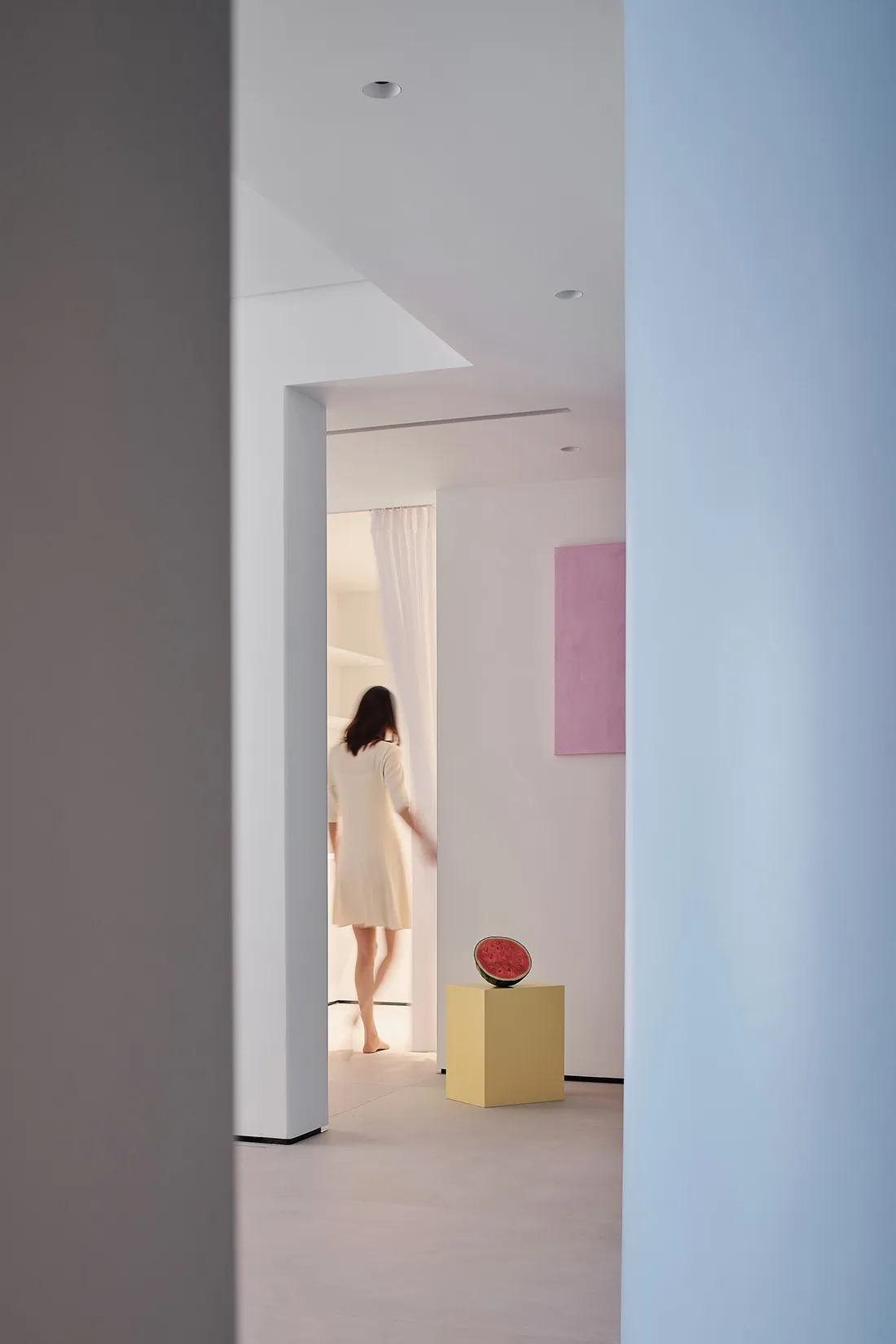 A minimalist white room with a pink wall and a yellow pedestal with a watermelon slice.