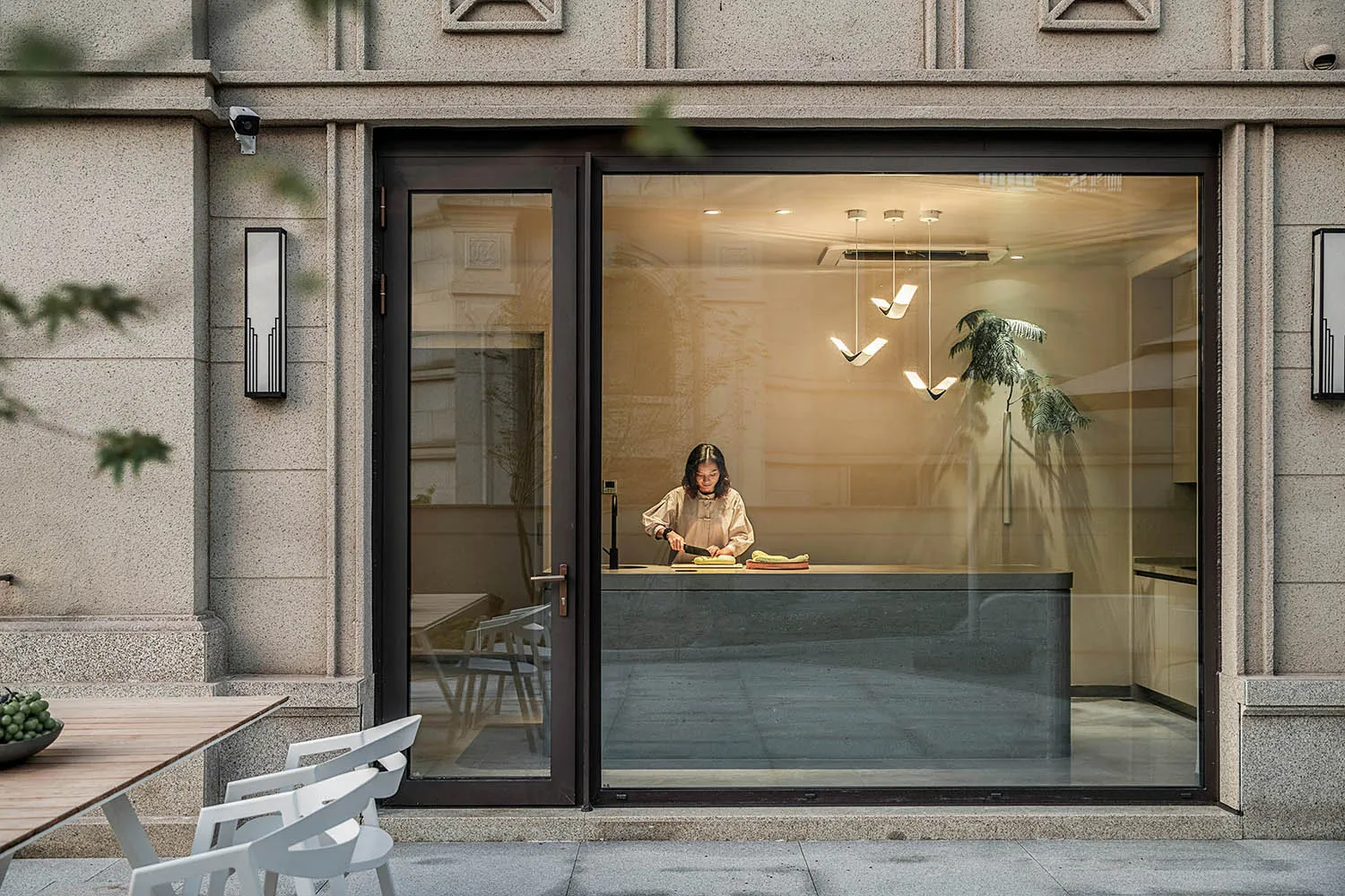 A modern kitchen with large glass walls, featuring a woman chopping vegetables on a counter.