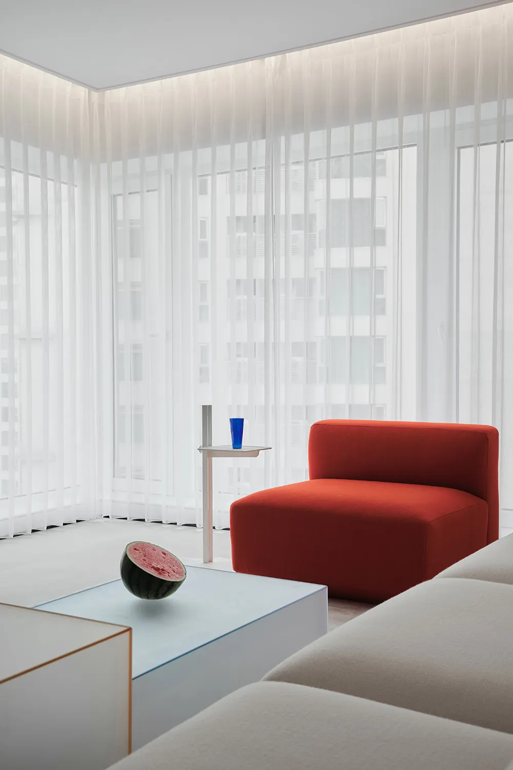 A modern living room with an orange upholstered sofa, white curtains, and a glass table with a half-cut watermelon and blue c