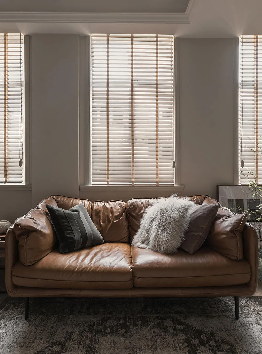 A modern leather sofa adorned with throw pillows and placed in front of a window with wood blinds.
