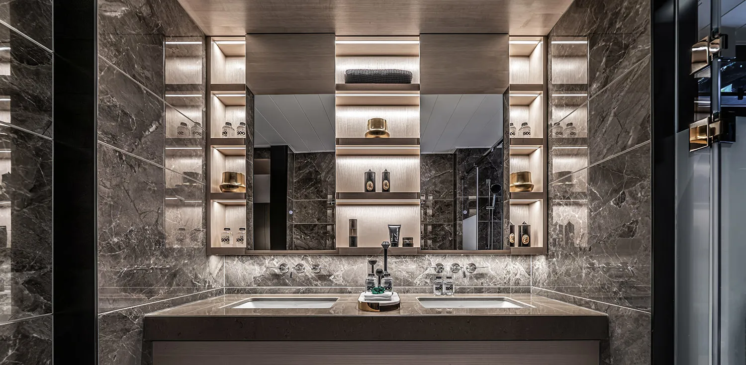 Modern luxury bathroom featuring marble walls and countertops with a double sink and illuminated shelves.