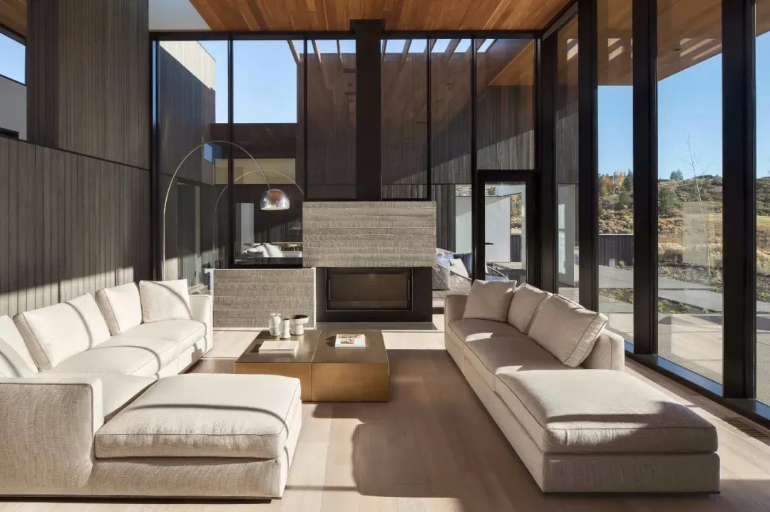 A modern living room with white sofas, a gold coffee table, and a fireplace, featuring wood paneling, metal accents, and glas