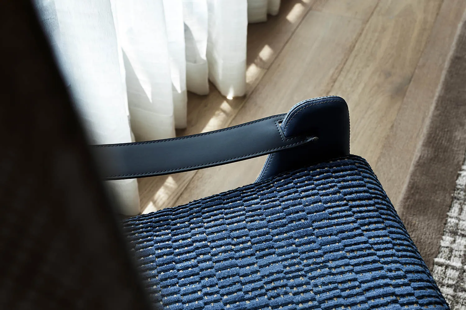 A close-up of a blue textured cloth leather chair with a wooden floor and a white curtain in the background.