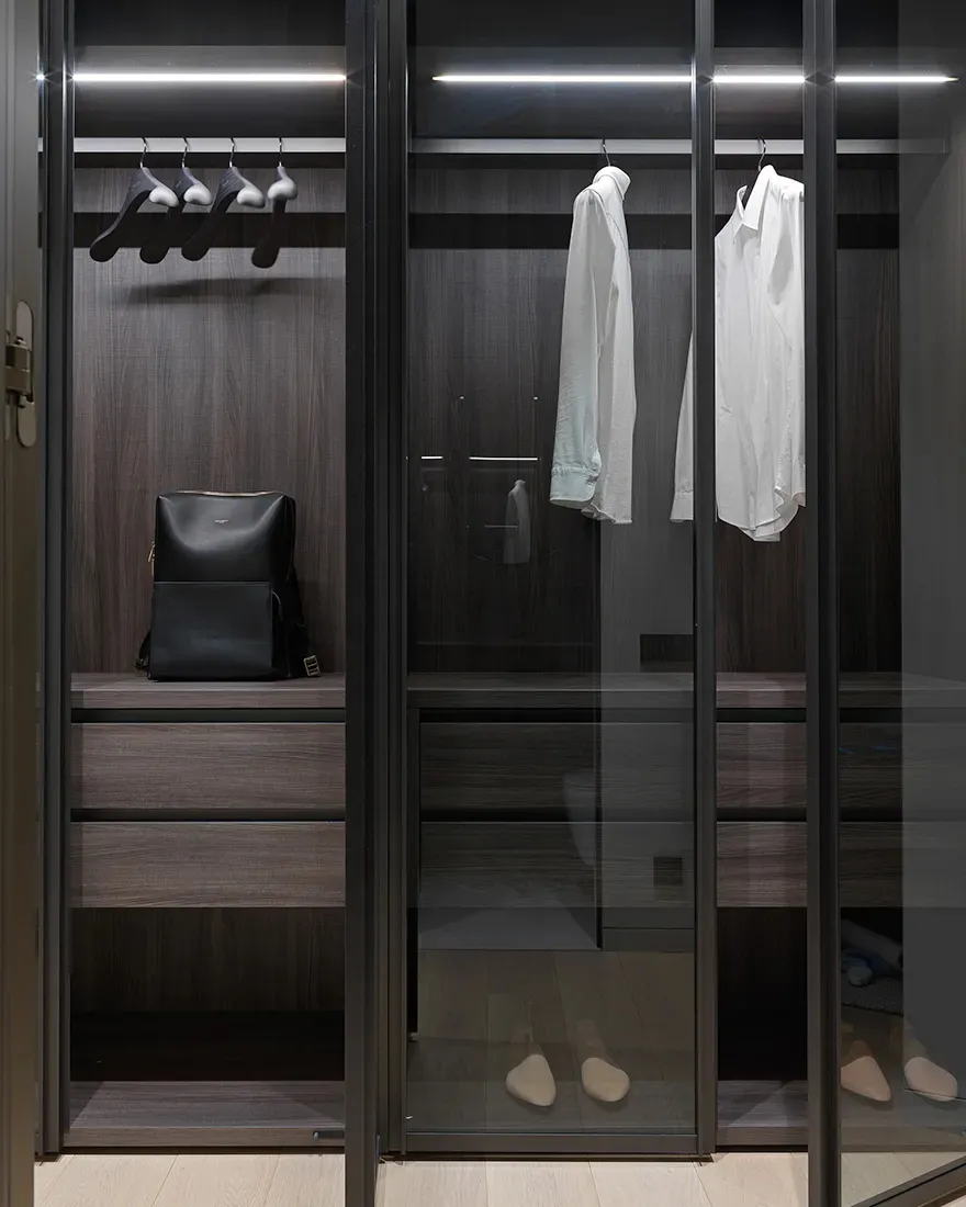 Modern dark wood metallic closet with glass doors, hanging clothes, and shoe rack.