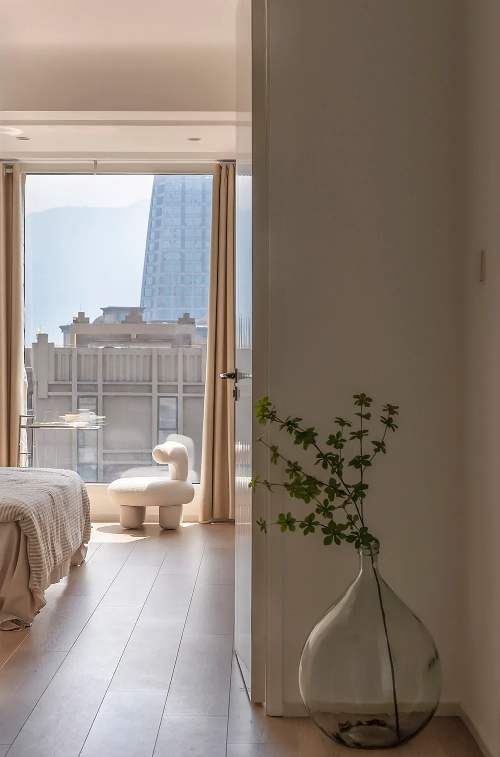 A modern residential interior with a white floor, beige curtains, and a glass vase with green plants.