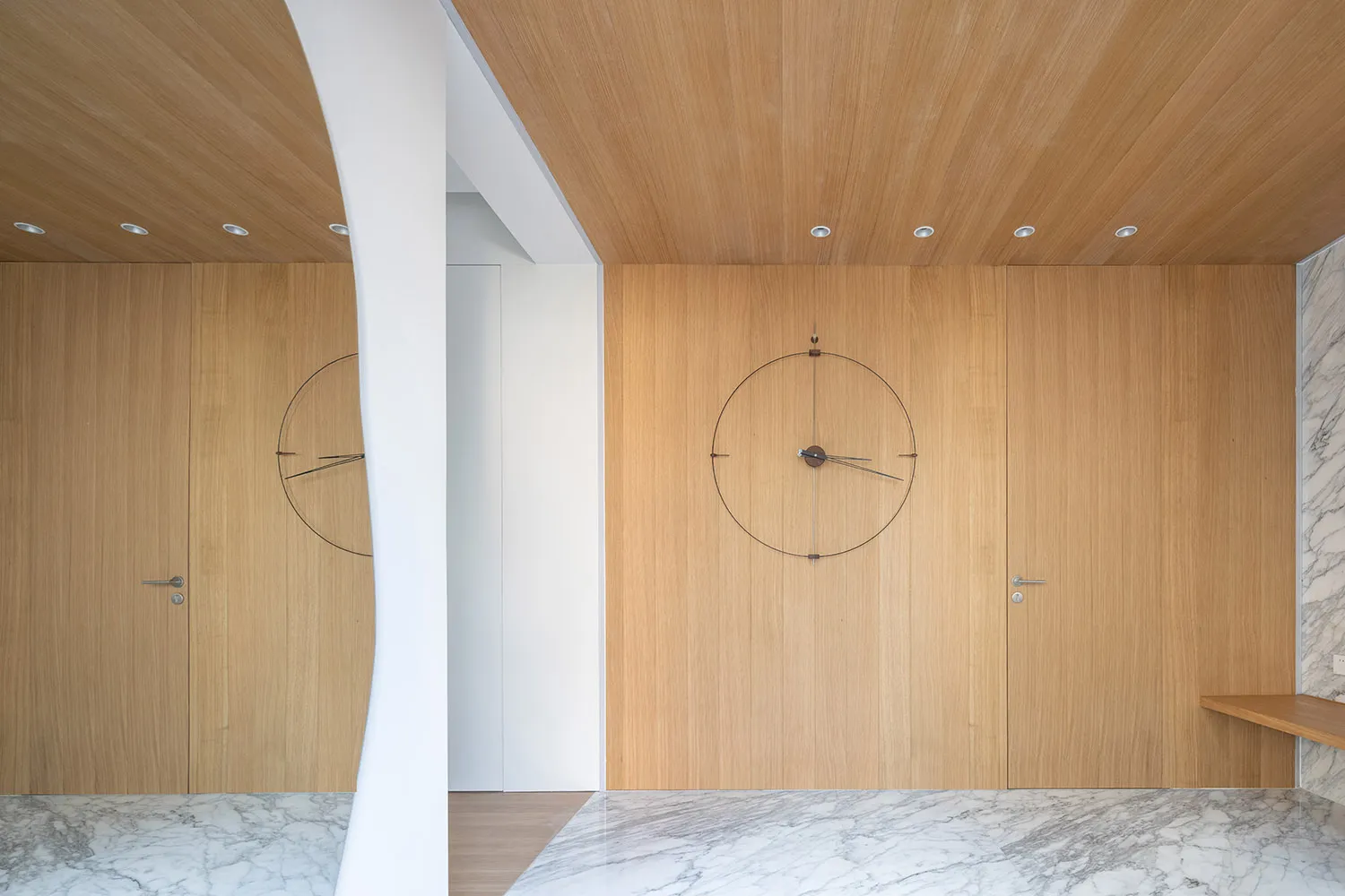 A modern wooden door with a large circular clock embedded in it, situated in a minimalist interior space with white walls and