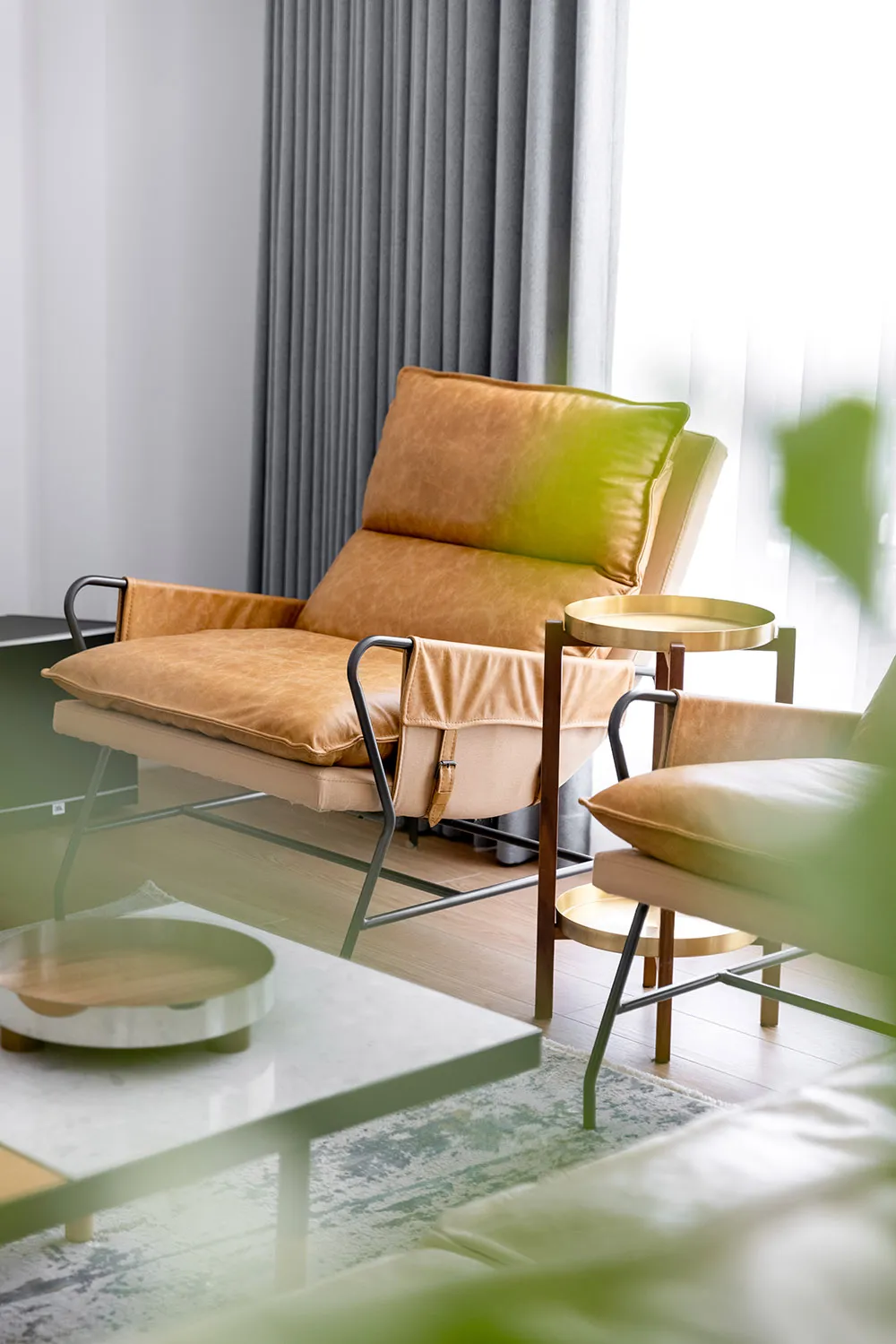 A minimalist living room featuring a leather armchair and sofa, with a glass coffee table and a green plant in the foreground