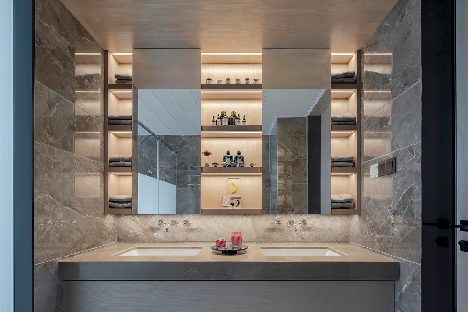 A minimalist bathroom with marble walls and floors, featuring two sinks and built-in shelves.