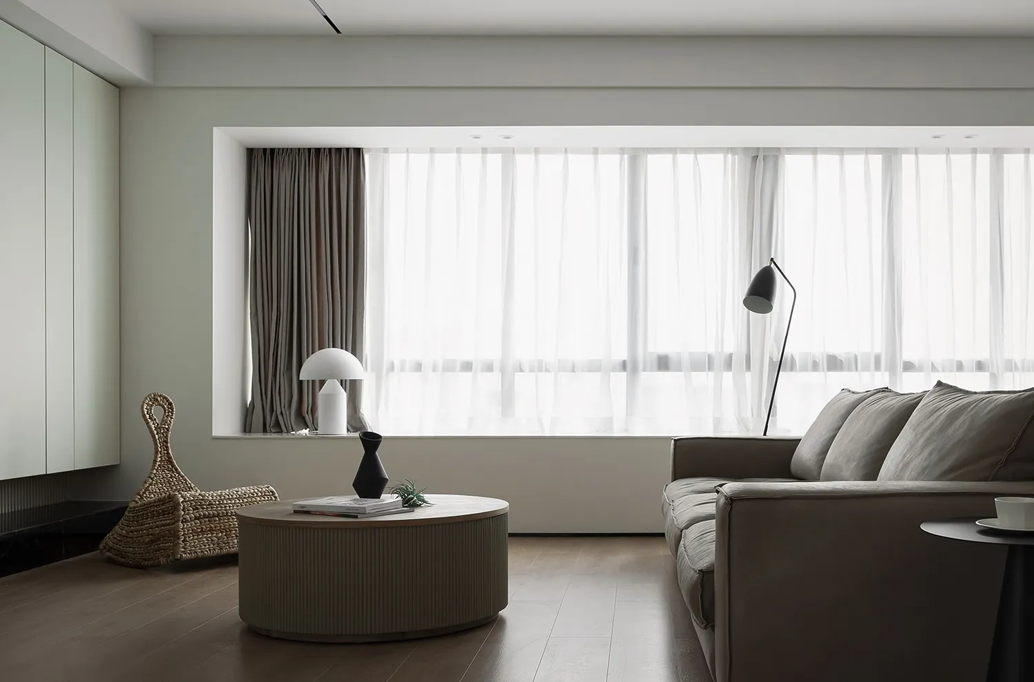 A modern living room with a light wood floor and a large floor-to-ceiling window with sheer curtains.