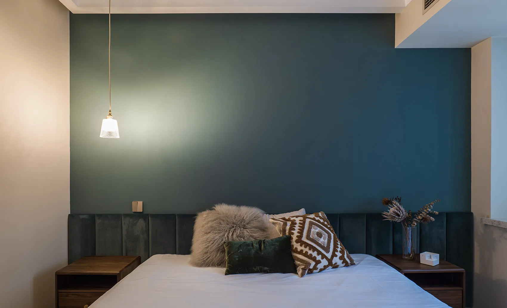 A minimalist bedroom featuring a bed with a velvet headboard, decorated with various pillows and a fur blanket, illuminated b