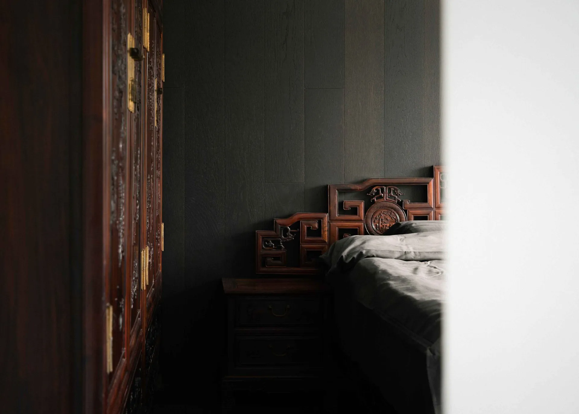 A minimalist bedroom with a wooden headboard, neatly made bed, and dark wooden furniture against a dark grey wall.