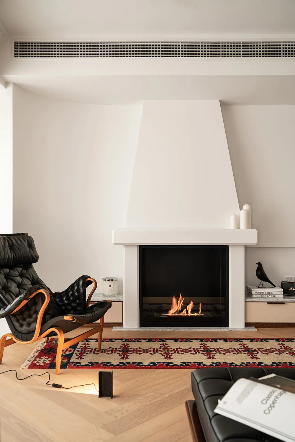 A minimalist living room with a fireplace, hardwood floors, a black leather chair, and a patterned rug.