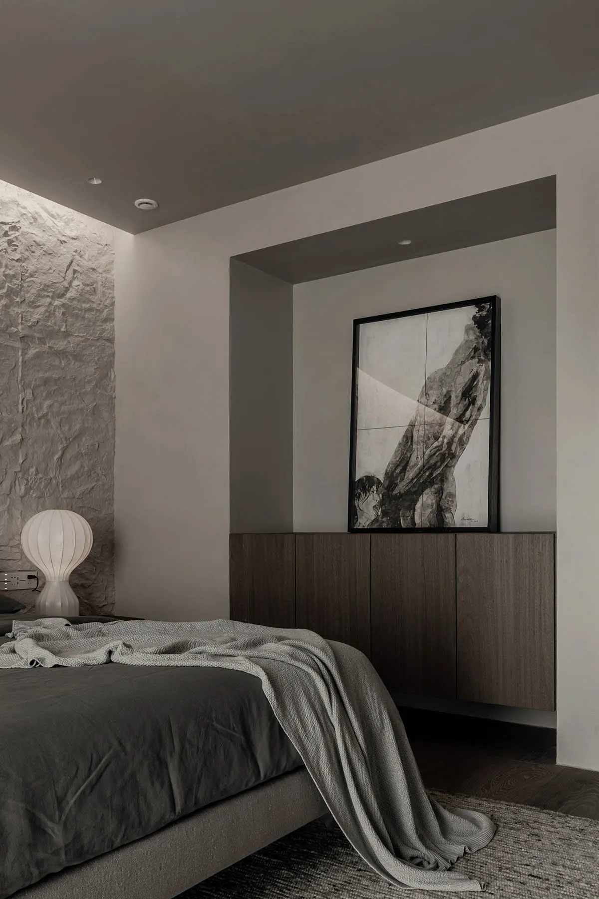 A minimalist bedroom with a textured stone wall, a bed with gray bedding, and a framed artwork.