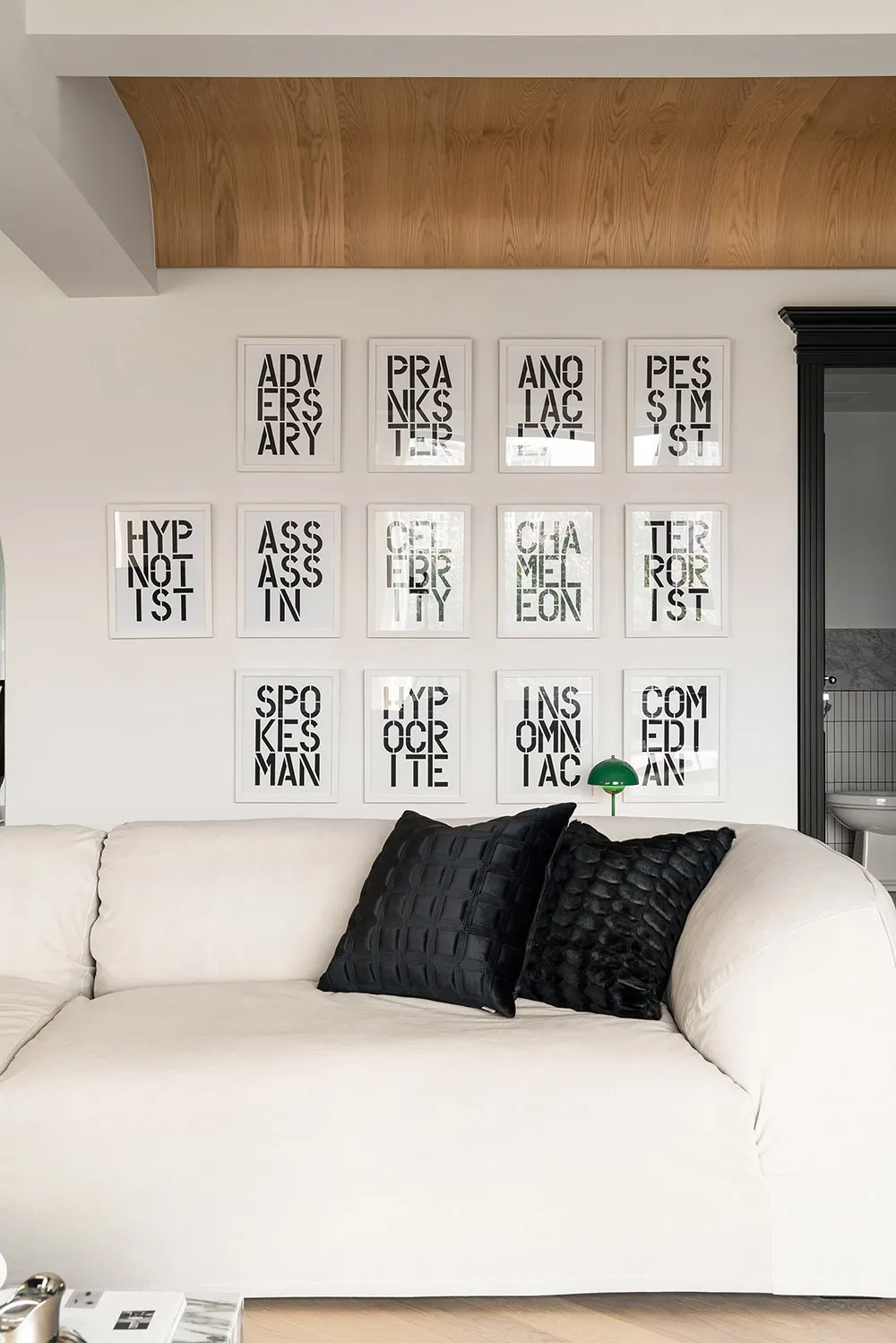 A minimalist living room with a white couch adorned with black cushions, set against a white wall adorned with framed words.