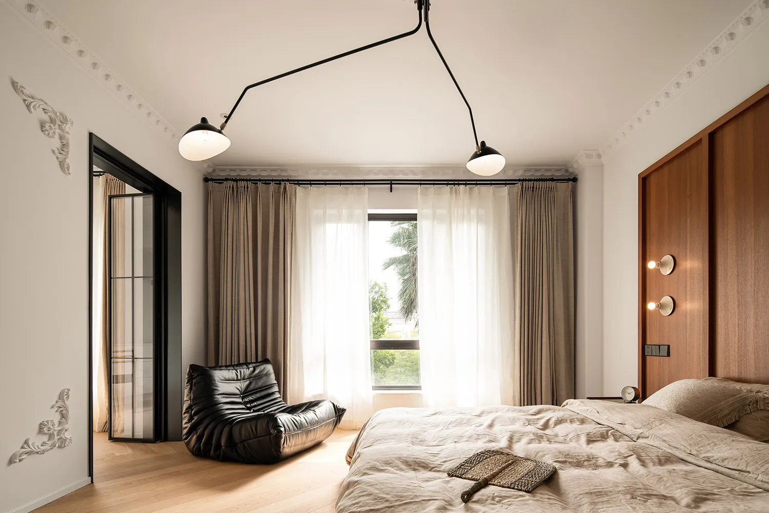 A minimalist bedroom with a wooden floor, a black leather bean bag, a bed with a wooden headboard, and natural light coming t