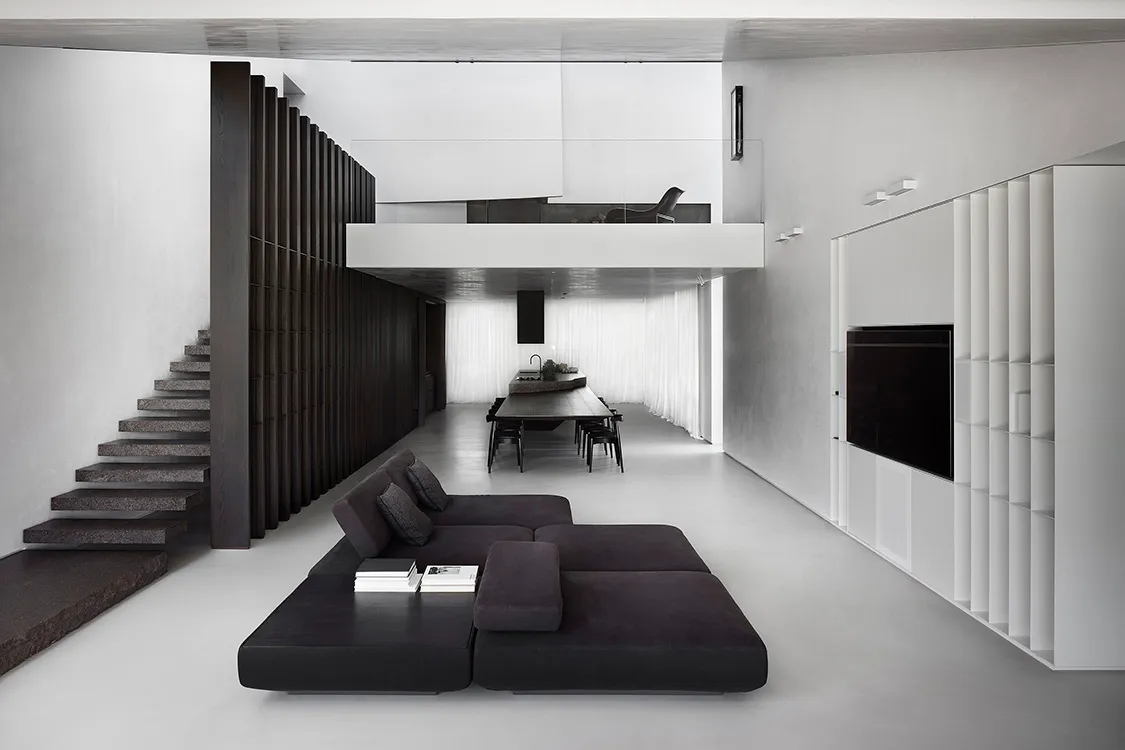 Modern minimalist living room with concrete floor, black sofa, and a raised dining area.