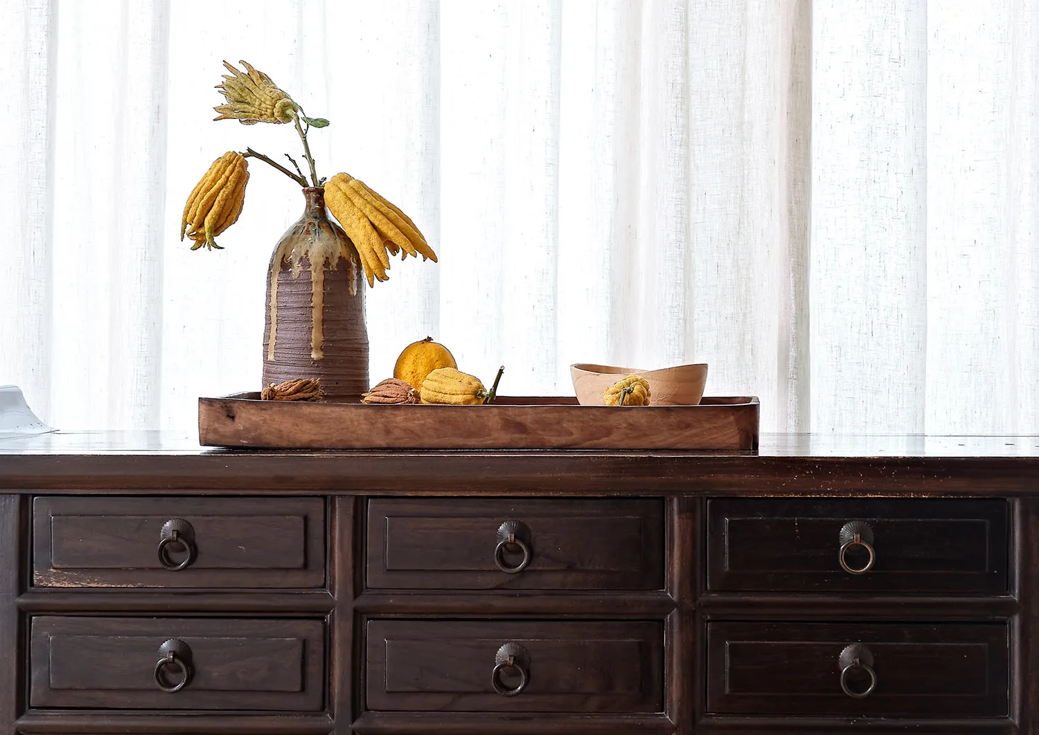 A wooden tray with a ceramic vase and fabric-covered items on a wooden dresser.