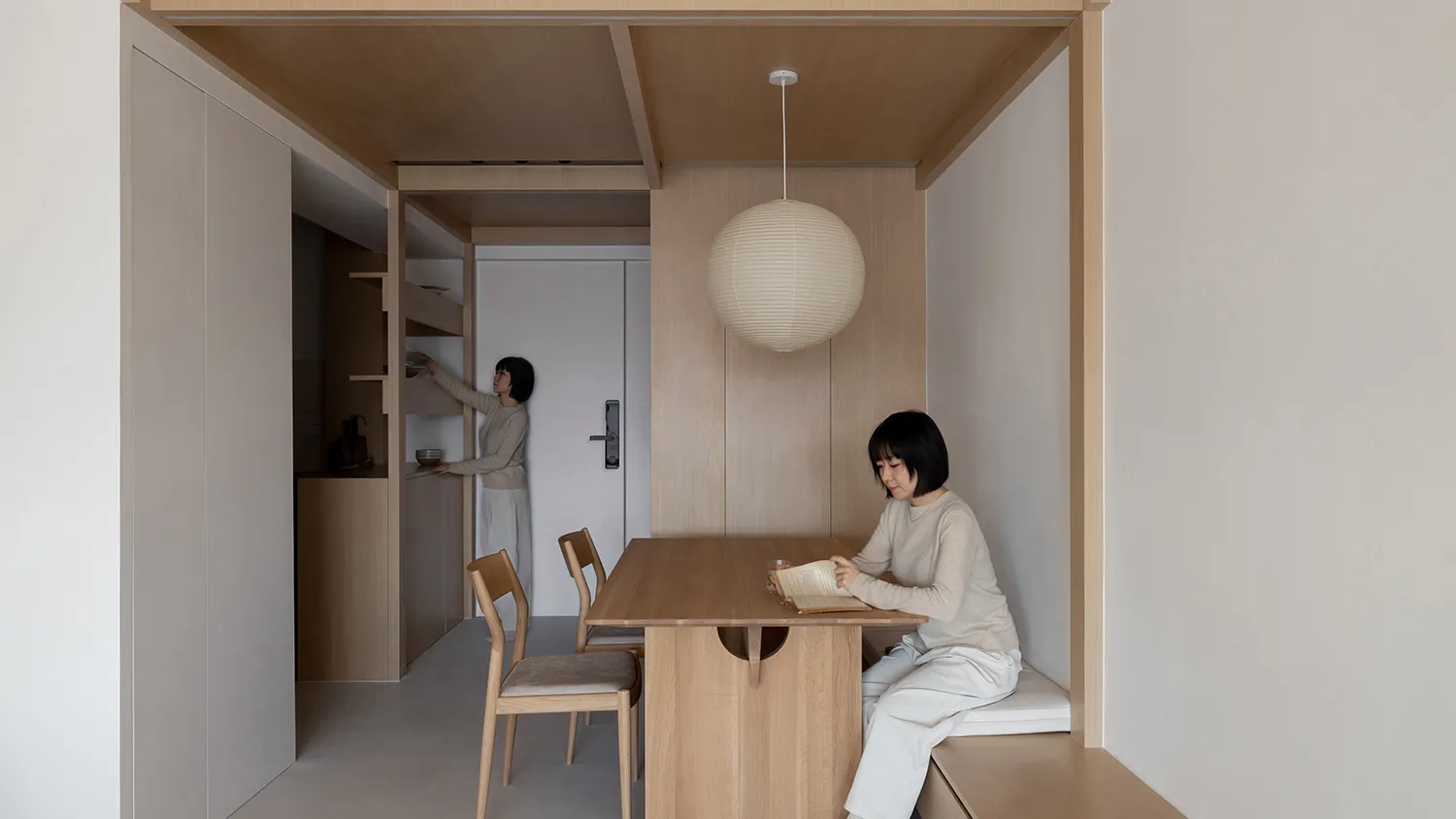Person sitting in a minimalist dining area with wooden furniture and a round light fixture.