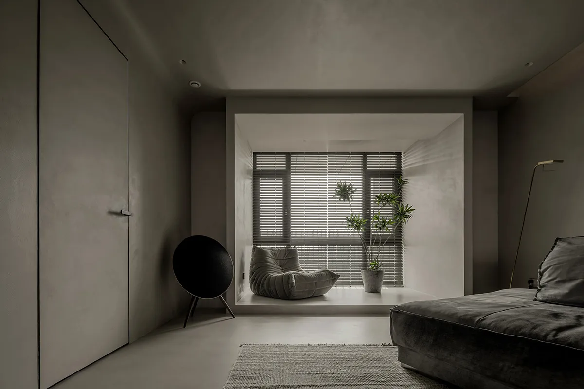 A minimalist living room with concrete walls and a sofa, a window with blinds, and a speaker.