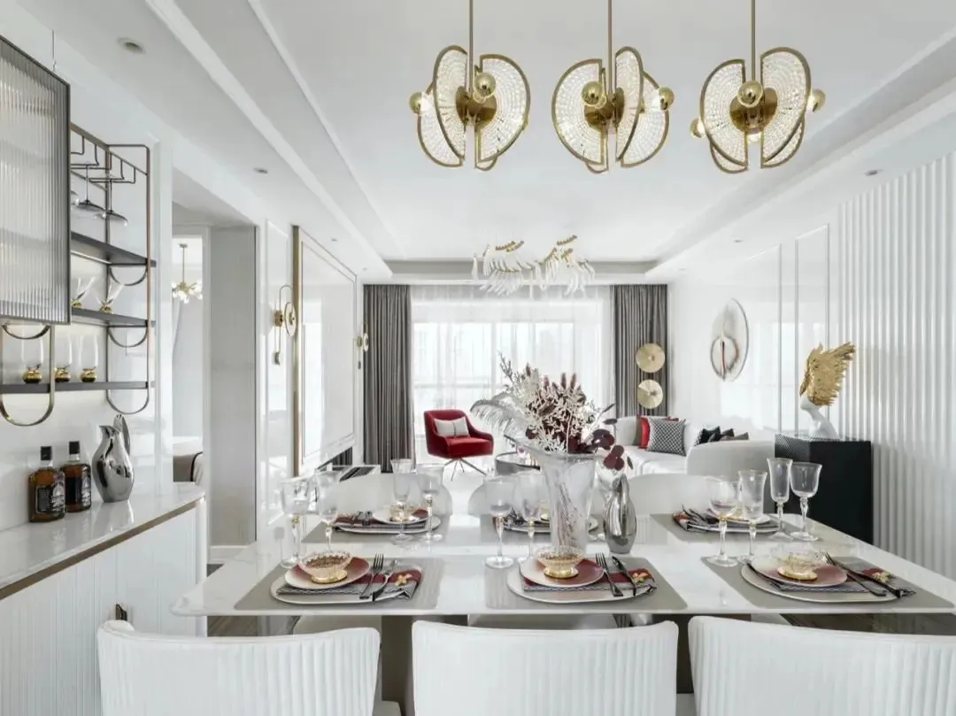 Modern dining room with gold and white decor, white table, red accent chairs, crystal chandelier, and metallic accents.