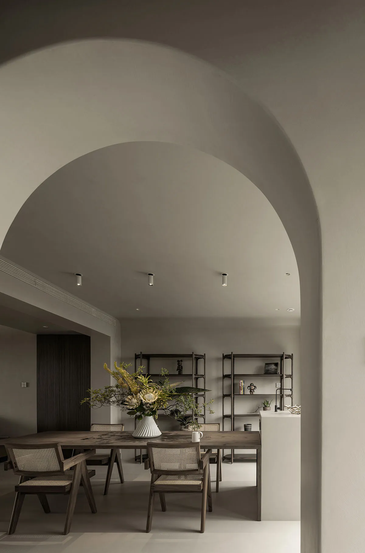 A modern minimalist dining room featuring a wooden table and chairs, with a large vase of flowers and bookshelves in the back