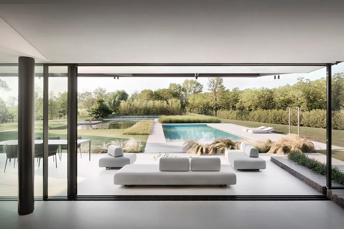 Modern residential living area with glass walls overlooking a pool surrounded by greenery.