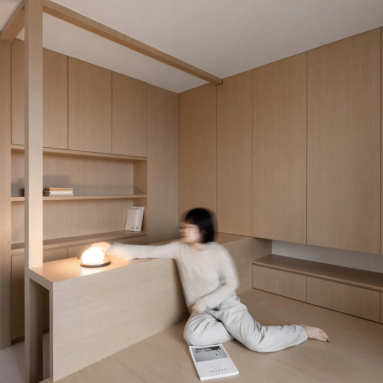 A minimalist residential interior with warm wood materials and a person adjusting a table lamp.