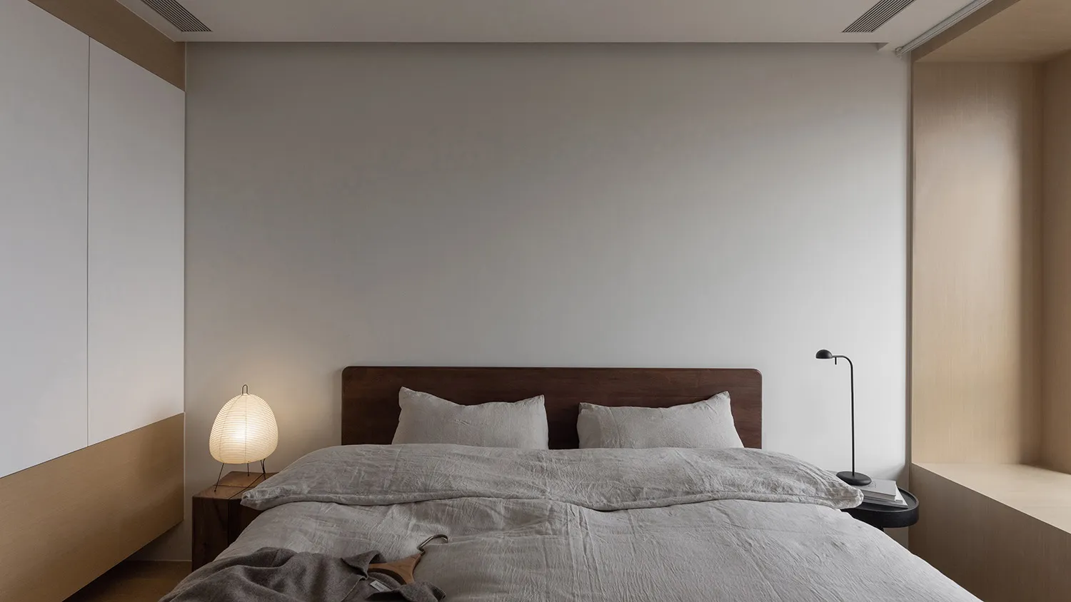 A minimalist bedroom with a bed, nightstand, and lamp, featuring neutral-toned bedding and decor.
