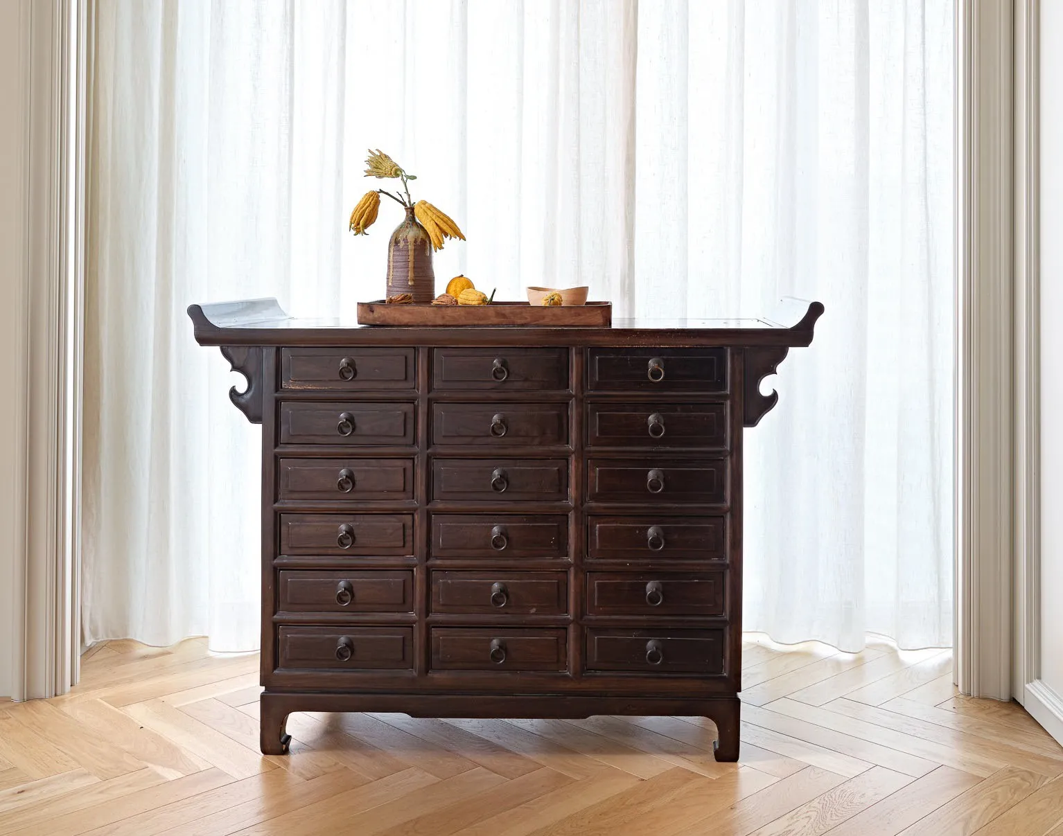 Dark wooden cabinet with numerous drawers and a vase with dried flowers and fruits on top.