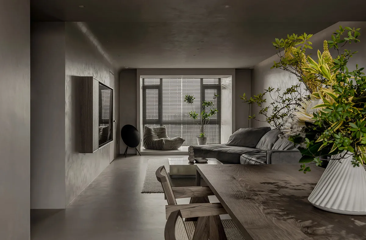 A modern, minimalist living room featuring a concrete floor, muted-colored furniture, and a large window with greenery.